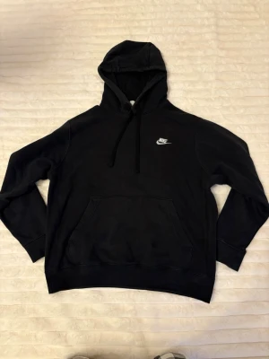 Svart Nike hoodie med logga - Svart hoodie från Nike med vit broderad logga på bröstet. Klassisk modell med huva och dragsnören samt stor magficka framtill. Mjuk och skön bomullsblandning, perfekt för en avslappnad streetstyle-look.