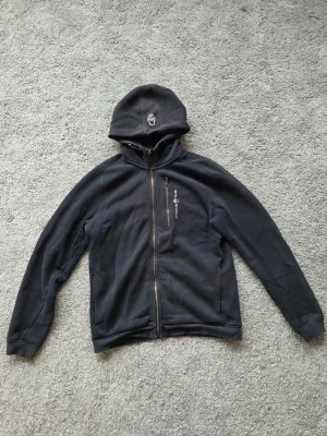 Svart hoodie från Sail Racing - Svart hoodie från Sail Racing med dragkedja framtill och en bröstficka med logga. Huva med broderad logotyp och två sidofickor. Tillverkad i mjukt material som känns skönt mot huden. Perfekt för dig som gillar en clean och sportig stil.