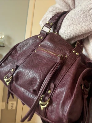 Vinröd handväska med nitar och dragkedja - Snygg vinröd handväska i skinnimitation med flätade handtag, guldfärgade metalldetaljer och dekorativa nitar. Väskan har en dragkedjeficka framtill och långa tofsar som detaljer. Perfekt storlek för det viktigaste och ger en cool vibe till din outfit. Från bershka, köpt i Spanien.