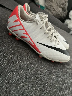 Vita Nike fotbollsskor med röd detalj - Säljer ett par snygga vita Nike fotbollsskor med stora svarta swoosh-loggor och orange/röda detaljer på hälen och sulan. Skorna har snörning och är tillverkade i ett slitstarkt syntetmaterial som ger bra bollkontroll på planen. Perfekta för dig som vill sticka ut på matchen!