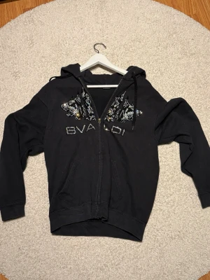 Svart hoodie från Byaloi med vargmotiv - Svart hoodie med dragkedja från Byaloi. På bröstet finns ett detaljerat vargmotiv i paljetter och texten BYALOI. Hoodien har huva med svarta snören och två fickor framtill. Perfekt för dig som gillar streetwear och vill sticka ut.