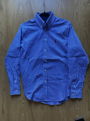 Blårutig skjorta från Polo Ralph Lauren - Snygg blårutig skjorta från Polo Ralph Lauren med klassisk button-down krage och broderad röd logga på bröstet. Skjortan har lång ärm och vita knappar framtill. Tillverkad i mjuk bomull som känns skön mot huden.