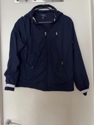 Marinblå vindjacka från Polo Ralph Lauren - Snygg marinblå vindjacka från Polo Ralph Lauren med vit logga på bröstet. Jackan har dragkedja framtill, två fickor med dragkedja och vitrandiga muddar vid ärmslut och huva. Tillverkad i lätt och tåligt material, perfekt för blåsiga dagar.