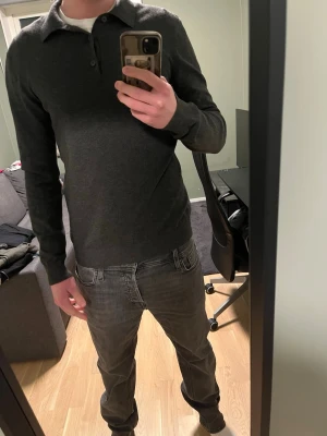 Långärmad pikétröja - Säljer en stilren mörkgrå långärmad pikétröja med krage och knappar framtill. Tröjan har en smal passform och är tillverkad i ett mjukt, tunt material som känns skönt mot huden. Perfekt för dig som gillar en clean och enkel look.