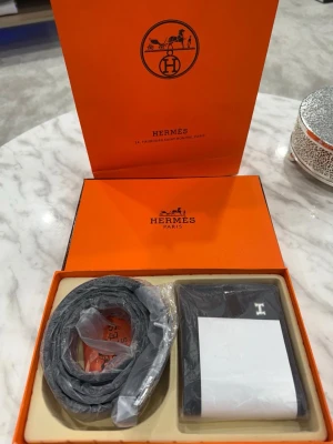 Exklusivt läderset med bälten och plånbok  - Ett stilrent lyxset i svart läder med eleganta silverdetaljer. Det består av ett klassiskt bälte med ett karaktäristiskt H-spänne, en matchande plånbok och exklusiva orangea originalförpackningar som ramar in helheten. 