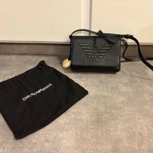 Hand/axelremsväska - Helt ny handväska med ställbara axelband. Köpt för flera år sedan på Zalandoo. Finns 2 små skav längst in på axelbandet på vardera sida. Då väskan bara legat i påsen och märkena är precis där det legat mot ’locket’ på väskan. Dock inget som syns och syns mer på bild än i verkligheten (se 2 sista bilderna) Stängs med metall. Bricka bredvid med text ’Emoprio Armani’. Ett större fack fram, samt ett litet. Bak är det 2 fack, varav ena stängs med liten knapp. Finns fler bilder om man vill ha. 