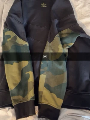Adidas hoodie med camoärmar - Säljer en svart Adidas hoodie med kamouflagemönstrade ärmar och klassisk Adidas-logga på bröstet. Hoodien har en avslappnad passform och är tillverkad i mjukt material. Perfekt för dig som gillar streetwear och vill sticka ut lite extra.