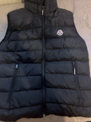 Svart dunväst från Moncler - Snygg svart dunväst från Moncler med klassisk logga på bröstet och två dragkedjeförsedda fickor framtill. Västen har hög krage och quiltad design, samt dragkedja hela vägen upp. Perfekt för lager-på-lager och streetstyle.