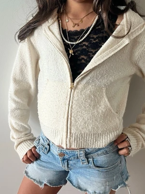 Krämvit hoodie med dragkedja - Mysig krämvit hoodie med dragkedja framtill och huva. Tillverkad i ett fluffigt, teddy-liknande material och har ribbade muddar vid ärmslut och nederkant. Perfekt för en chill och avslappnad stil.