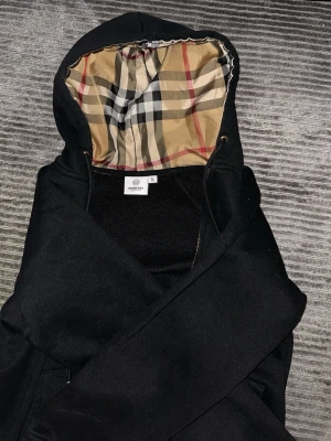 Svart hoodie från Burberry - Säljer en svart hoodie från Burberry med dragkedja och klassiskt rutigt foder i huvan. Hoodien har justerbara snören och fickor framtill. Perfekt för dig som vill ha en stilren och lyxig look med ikoniskt mönster i huvan. DOCK står XL men det är helt fel det ska va S, priset kan variera beroende på pengarna jag får hör av er om ni har fler frågor.
