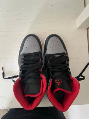 Air Jordan 1 Mid svart/grå/röd - Snygga Air Jordan 1 Mid sneakers i svart och grått med röda detaljer och röd insida. Klassisk Nike Swoosh på sidan och perforerad tåbox. Skorna har högt skaft och svart snörning. Yttermaterial i läder och textil, sula i vitt och rött.