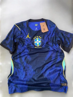 Blå Brasilien original tröja Nike Jordan - Snygg blå matchtröja från Brasilien med gul Nike Jordan-logga och CBF-emblem på bröstet. Tröjan har ett mönstrat tryck i olika blå nyanser, gula detaljer vid axlar och sidor samt rund hals. Tillverkad i lätt och ventilerande funktionsmaterial.