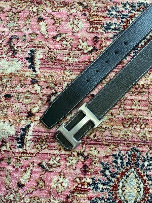 Svart bälte med H-spänne - Säljer ett svart skärp i skinn med ett stort H-format spänne i silverfärg. Skärpet har vit kontrastsöm längs kanterna och flera hål för justerbar passform. Perfekt accessoar för att lyfta vilken outfit som helst. Bältet är 105cm men finns även i 100cm! Priset går absolut att diskuteras! 