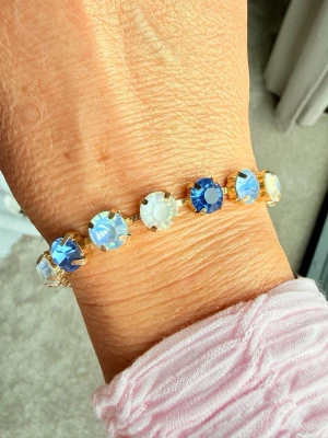 Armband med blå och vita stenar  - Snyggt armband med 8mm stora runda  stenar i sapphirblått och vitt, infattade i guldfärgad metall. Armbandet har justerbar kedja och gnistrar superfint i ljuset. Perfekt accessoar för att lyfta din outfit med lite extra färg och bling. Bild 4 är en inspirationsbild hur du kan kombinera med egna smycken från tex Svedbom. Bild 5 är ett urval av färger som finns! 