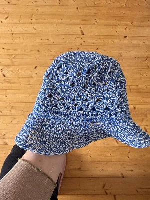 Blåvit virkad bucket hat - Säljer en handvirkad bucket hat i blått och vitt garn. Hatten har ett snyggt mönster med blommiga detaljer och bred brätte. Perfekt accessoar för att sticka ut i mängden och ge din outfit en cool touch.
