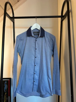 Blå skjorta med mörkblå krage - Snygg blå skjorta  med kontrasterande mörkblå krage och svarta knappar. Skjortan har lång ärm och klassisk passform, perfekt för en stilren look. Tillverkad i bomull för en bekväm känsla.
