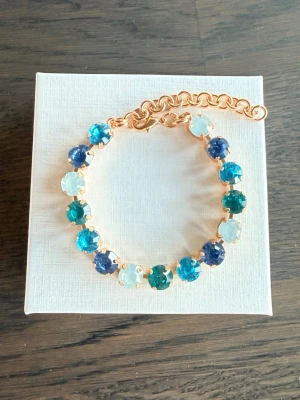 Armband med blå/turkosa stenar  - Snyggt armband med 8mm stora glittriga stenar i olika blå och turkosa nyanser, infattade i guldfärgad metall. Justerbar kedja för perfekt passform. Perfekt accessoar för att lyfta din outfit med lite färg och glans.