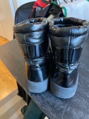Lejon Svarta moon boots med snörning stl 34 - Säljer ett par klassiska svarta moon boots med snörning framtill och vadderad, quiltad look. Skorna har en glansig yta och tjock sula, perfekta för kalla dagar. Stilsäkra och bekväma för vintern!
