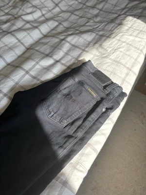 Nudie jeans  - Säljer ett par svarta jeans från Nudie Jeans med klassisk femficksdesign och smal passform. Jeansen har diskreta detaljer och är tillverkade i stretchigt denim för extra komfort. Perfekta för en stilren och enkel look.