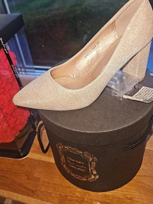 Glittriga champagnefärgade pumps - Snygga pumps i champagnefärgat glittrigt material med spetsig tå och bred klack. Skorna har en elegant och modern design som verkligen sticker ut. Perfekta för dig som vill ha något extra till outfiten. Märket är inte synligt. Nypris 399kr köpta i Danmark 