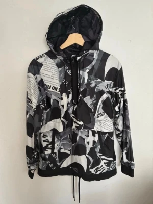 Nike vindjacka med svartvit print street cool - Nike vindjacka med grafiskt svartvitt mönster och textdetaljer. Jackan har huva med dragsko, halv dragkedja framtill och elastiska muddar. Lätt material, perfekt för streetwear och sportig stil.
