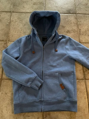 Blå hoodie med dragkedja från Medicine - Blå hoodie från Medicine i storlek S med dragkedja framtill, huva med dragsko och fickor på sidorna. Snygga detaljer i brunt läderimitation vid snören och ficka. Tillverkad i mjukt bomullsmaterial för en skön känsla.