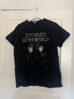 Svart Avenged Sevenfold t-shirt M - Avenged Sevenfold merch. T-shirten har ett stort motiv med dödskalle och krona, samt bandets logga i grått. Storlek M
