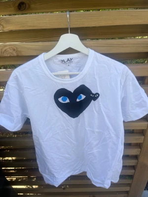 Vit Comme des Garçons Play t-shirt - Vit t-shirt från Comme des Garçons Play med det ikoniska svarta hjärtat med blå ögon tryckt på bröstet. Klassisk rund hals och korta ärmar. Tillverkad i mjuk bomull för en skön känsla och stilren look.
