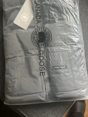 Grå dunväst från Canada Goose - Snygg grå dunväst från Canada Goose med två stora fickor framtill och diskret logotyp på bröstet. Perfekt för lager-på-lager och håller dig varm med sin klassiska, stilrena design. !!Kvitton kastades vid ankomsten skicka offer vid intresse!!