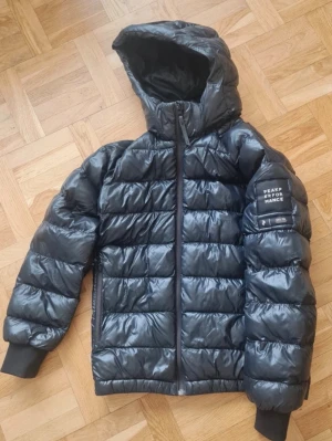 Svart dunjacka från Peak Performance - Snygg svart dunjacka från Peak Performance med glansig finish och diskret logga på bröstet. Jackan har dragkedja framtill och quiltad design som håller värmen under kalla dagar. Perfekt för dig som vill ha både stil och funktion i vinter nåra små hål lite här o där o ganska sliten