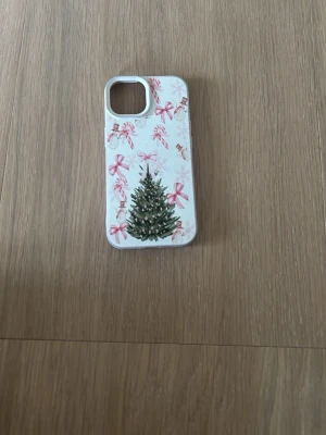 Mobilskal med julgran och rosetter - Snyggt mobilskal med vit bas, dekorerat med en grön julgran och rosa rosetter samt små julmotiv. Perfekt för dig som vill ha en festlig look på mobilen under vintern. iPhone 15
