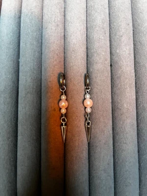 Hängande örhängen med pärlor och spikes - Säljer ett par coola örhängen med små rosa pärlor och metallspikar. Örhängena har en silverfärgad metall och en modern design med två pärlor på rad och en spetsig detalj längst ner. Perfekta för dig som gillar edgy accessoarer.