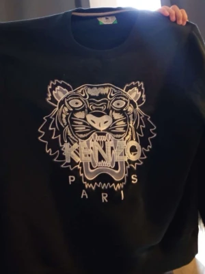 Svart stickad Kenzo-tröja med tiger - Cool svart stickad tröja från Kenzo med ikoniskt tigerbroderi och texten 'KENZO PARIS' på bröstet. På ärmen finns en patch med 'JUNGLE KENZO PARIS' och tiger. Perfekt för dig som gillar streetstyle och statementplagg.