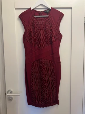 Vinröd kort klänning från Lipsy London - Vinröd kort klänning från BCBGMAXAZRIA med snygga spetsdetaljer och geometriskt mönster framtill. Ärmlös design med figurnära passform och dragkedja i ryggen. Perfekt för dig som vill sticka ut med en elegant look.