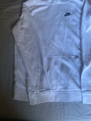 Vit Nike hoodie med svart logga - Snygg vit hoodie från Nike med svart broderad logga på bröstet. Klassisk känguruficka framtill och huva. Perfekt för en avslappnad och sportig stil. Mjuk bomullskänsla och ribbade muddar vid ärmslut och nederkant.