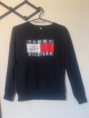 Svart sweatshirt Tommy Hilfiger - Svart sweatshirt från Tommy Hilfiger med stort tryck på bröstet i blått, vitt och rött. Klassisk rund halsringning och långa ärmar. Perfekt för en avslappnad och stilren look.