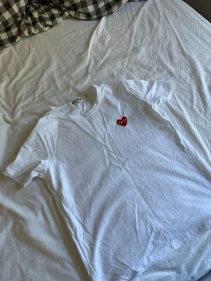 Vit t-shirt från Comme des Garçons Play - Vit t-shirt från Comme des Garçons Play med det ikoniska röda hjärtat med ögon broderat på bröstet. Klassisk rund hals och korta ärmar. Tillverkad i mjuk bomull som känns skön mot huden. Perfekt för en clean och stilren look.