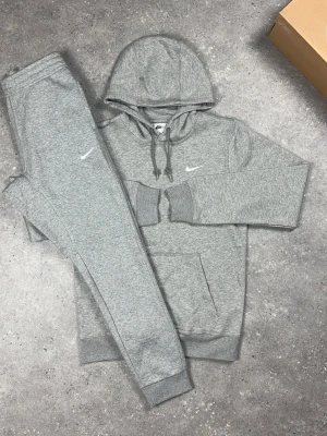 Grått Nike loungewear set - Snyggt och bekvämt grått loungewear set från Nike med matchande hoodie och byxor. Hoodien har dragsko och magficka, byxorna har resår i midjan och smal passform. Båda plaggen har Nike-logga broderad i vitt. Perfekt för chill dagar hemma.