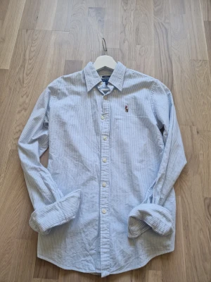 Ralph Lauren skjorta - Klassisk blårandig skjorta från Ralph Lauren i slim fit-modell. Skjortan har långa ärmar, knäppning framtill och den ikoniska broderade loggan på bröstet. Tillverkad i ett mjukt bomullstyg som känns skönt mot huden. Perfekt för en stilren och avslappnad look.