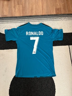 Real Madrid Ronaldo 7 bortatröja - Säljer en blå Real Madrid bortatröja från Adidas med Ronaldo 7 på ryggen. Tröjan har Champions League-patchar på ärmarna, klubbmärke och sponsortryck framtill. Tillverkad i lätt och ventilerande material, perfekt för fotbollsträning eller match.dm vid intresse