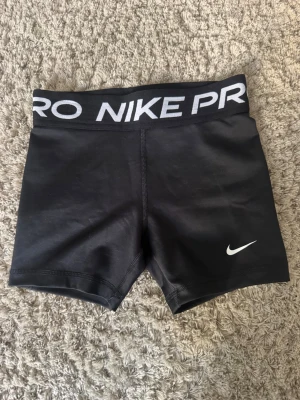 Svarta Nike Pro träningstights-shorts - Svarta Nike Pro shorts med bred resår i midjan där det står 'NIKE PRO' i vitt. Tight passform och korta ben, perfekt för träning. Materialet är stretchigt och känns svalt mot huden. Klassisk Nike-logga framtill på benet. Aldrig använda. Lite osäker vilken storlek men skulle säga xs.