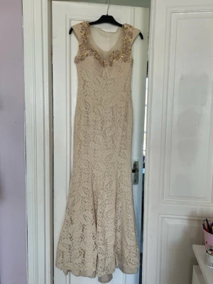 Beige spetsklänning med pärldetaljer - Elegant lång beige klänning med spets och v-ringning. Klänningen har vackra pärldetaljer och blommor vid axlarna och halsringningen. Ärmlös modell med figurnära passform och lätt utsvängd nederdel. Perfekt för speciella tillfällen.