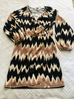 Poppydress  klänning från indiska helt ny - Snygg kort omlottklänning med svart, beige och vit zigzag-mönster. Klänningen har långa ärmar med resår vid handlederna och markerad midja. Perfekt för dig som gillar unika mönster och en trendig look.