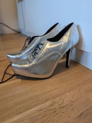 Silvriga pumps med snörning och platå - Snygga pumps i metallic-silver med hög klack och platåsula. Skorna har snörning framtill och en cool, modern design som sticker ut. Perfekta för dig som vill ha något unikt och trendigt. Materialet är glansigt och ger en futuristisk vibe.Finns någon skada på klacken se bild.Dessa är enbart provade.
