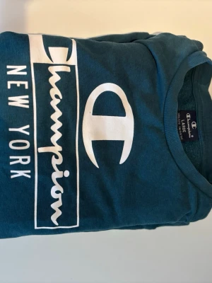 Blå Champion sweatshirt New York Large kids size (154cm) - Snygg blå sweatshirt från Champion med stort vitt tryck på bröstet där det står 'Champion New York'. Tröjan har rund halsringning och är tillverkad i mjukt material, perfekt för en avslappnad streetstyle.