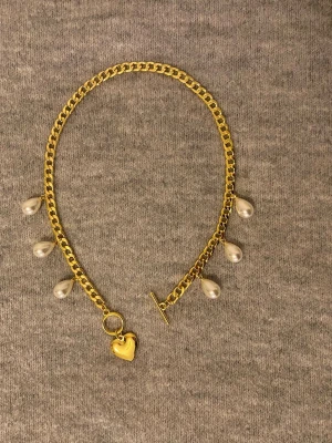 Guldfärgat halsband med pärlor och hjärta - Snyggt guldfärgat halsband med kedja, dekorerat med droppformade pärlor och ett litet hjärthänge. Halsbandet har en toggle-låsning och en trendig design som sticker ut. Perfekt accessoar för att lyfta din outfit.