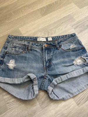 Blå jeansshorts med slitningar - Snygga blå jeansshorts med slitna detaljer och upprullade benslut. Klassisk femficksmodell med knappgylf och ljus tvätt. Perfekta för sommaren och ger en avslappnad vibe.
