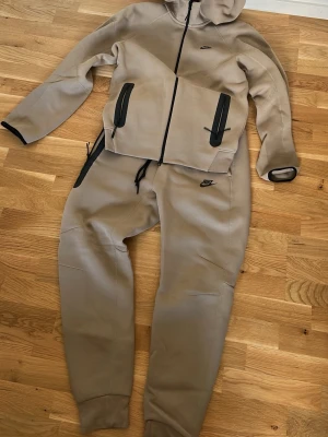 Beige mjukisbyxor från Nike - Snygga beige mjukisbyxor och tröja från Nike med svart logga på vänster sida. Byxorna har resår i midjan och vid bensluten samt praktiska fickor med dragkedja. Perfekta för en avslappnad och sportig stil. BYXORNA STORLEK M. TRÖJAN STORLEK S. 