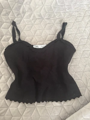 Svart ribbad linnetopp från Zara - Svart ribbad linnetopp från Zara med smala axelband och vågig kant nertill och upptill. Toppen är figurnära och har en enkel, stilren design som passar perfekt till jeans eller kjol.