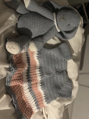 Handvirkad klänning i pastellfärger och en virkad kanin - Supersöt handvirkad babyklänning i ljusblått, vitt och ljusrosa. Koftan har kort ärm, knappar framtill och ett luftigt, vågigt mönster nedtill. Perfekt för dig som gillar unika och mysiga plagg till de minsta. Gjord på bomull och passar 5 månader till 1 år ungefär och en blå egen gjord kanin gjord på bomull, går bra att köpa en och en men båda tsm blir priset de står 